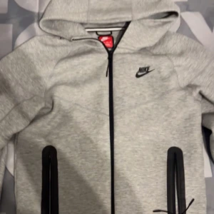 Grå hoodiejacka från Nike - Grå zip-hoodie från Nike med svart logga på bröstet. Jackan har huva, dragkedja framtill och två svarta dragkedjefickor. Perfekt för en sportig och avslappnad stil.