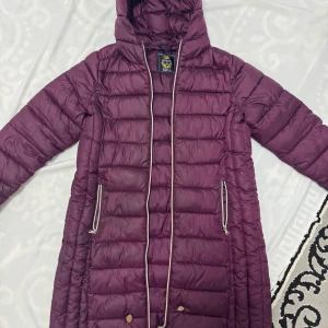 Vinröd lång dunjacka från My Wear Woman - Säljer en vinröd lång dunjacka från My Wear Woman i storlek L. Jackan har huva, dragkedja framtill och två fickor med dragkedja. Perfekt för kyliga dagar och har en klassisk pufferstil.