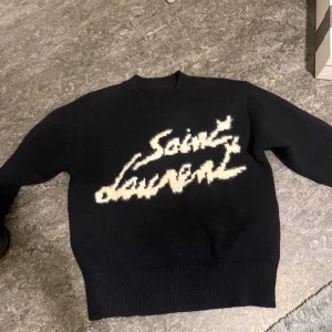  stickad tröja från Saint Laurent - Säljer en svart stickad tröja från Saint Laurent med vit, stor logotyp framtill. Tröjan har rund halsringning och långa ärmar. Perfekt för dig som vill ha en statement piece i garderoben.