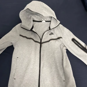 Grå zip-hoodie från Nike - Snygg grå hoodie från Nike med dragkedja och huva. Jackan har svarta detaljer, två fickor och en diskret Nike-logga på bröstet. Perfekt för en sportig och avslappnad stil.