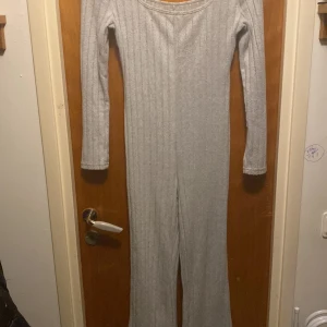 Ljusgrå ribbad jumpsuit med lång ärm - Säljer en ljusgrå ribbad jumpsuit med långa ärmar och utsvängda ben. Plagget har en bred, rundad ringning och är tillverkat i ett mjukt, stretchigt material som sitter skönt på kroppen.