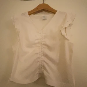 Vit blus  - Säljer en söt vit topp med v-ringning och volangdetaljer på axlarna. Toppen är ärmlös och har en rynkad söm framtill som ger en fin form. Använd Max två gånger. Tvättar innan jag skickar💕