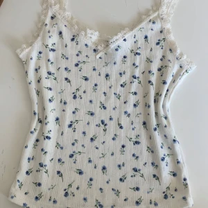 Coquette pointelle topp - Söt vit linnetopp från h&m med små blå blommor och spetsdetaljer längs urringningen. Toppen har smala axelband och en liten rosett framtill. Perfekt för varma dagar eller som ett lager under en kofta.