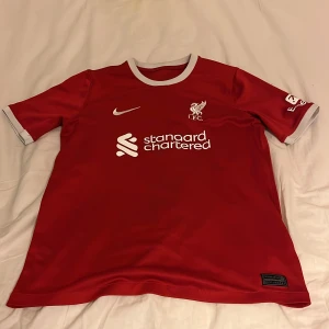 Liverpool FC röd fotbollströja från Nike - Säljer en röd Liverpool FC fotbollströja från Nike med  Dri-FIT. Officiell sponsorlogga och klubbmärke på bröstet. Perfekt för fans!