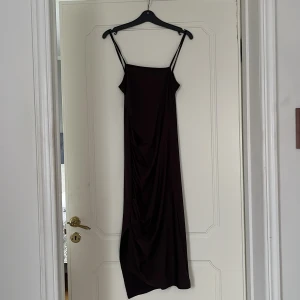 Mörkbrun långklänning med tunna axelband - Säljer en mörkbrun långklänning med smala axelband och draperad detalj på ena sidan. Klänningen sitter tight och är tillverkad i ett mjukt, stretchigt material som faller snyggt. Perfekt för dig som gillar minimalistisk stil.