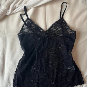 Svart spetslinne med rosett - Säljer ett svart linne i spets med smala axelband och en liten rosett framtill. Toppen har v-ringning och är figurnära med ett feminint uttryck. Perfekt att styla till jeans eller kjol. S-M