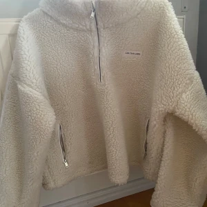 Vit teddyjacka med halv dragkedja från Lxa - Mysig vit teddyjacka med halv dragkedja framtill och två sidofickor med dragkedja. Jackan har hög krage och är tillverkad i fluffigt teddy-material. Perfekt för kyliga dagar och enkel att matcha med olika outfits.