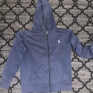 Blå hoodie från Polo Ralph Lauren - Säljer en blå hoodie från Polo Ralph Lauren med dragkedja och huva. Tröjan har två fickor framtill och en liten vit logga på bröstet. Perfekt för en avslappnad stil och enkel att matcha med jeans eller joggers. Sömmen gått upp lite på ena fickan. Köpt på vinted för 500kr
