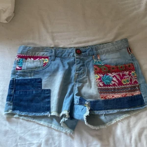 Desigual jeansshorts med broderi och fransar - Unika ljusblå jeansshorts med färgglada patchwork-detaljer och broderier framtill och baktill. Shortsen har råa fransade benslut och dekorativa mönsterband i midjan. Perfekta för dig som vill sticka ut med en kreativ och bohemisk stil.