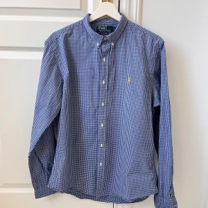 Blårutig skjorta från Polo Ralph Lauren - Säljer en stilren blårutig skjorta från Polo Ralph Lauren i slim fit. Skjortan har långa ärmar och knappar framtill. Den är broderad med det klassiska Polo Ralph Lauren-emblemet i gult på bröstet. Väldigt bra skick förutom lite missfärgning som du ser på sista bilden. Märks inte