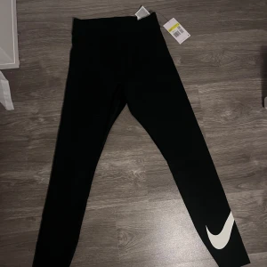 Svarta leggings med vit nike logga - Snygga svarta leggings från Nike med en stor vit logga på benet. De har en tight passform och hög midja, vilket ger en bekväm och stilren look. Perfekta för träning eller en avslappnad dag.