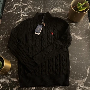 Svart stickad tröja från Polo Ralph Lauren - Säljer en stilren svart stickad tröja från Polo Ralph Lauren med kabelstickat mönster och en liten röd broderad logotyp på bröstet. Tröjan har en dragkedja vid halsen och långa ärmar. Perfekt för kyliga dagar!