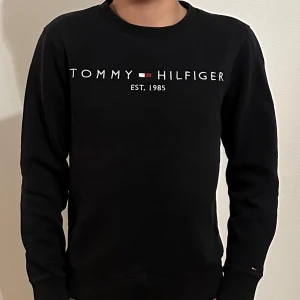 Svart tröja från Tommy Hilfiger - Snygg svart tröja från Tommy Hilfiger med deras ikoniska logga och text 'Est. 1985' på framsidan. Tröjan har långa ärmar och en klassisk rund halsringning. Perfekt för en stilren look.