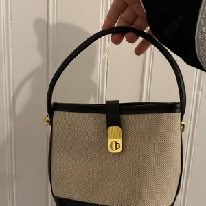 Beige och svart handväska med gulddetalj - Elegant handväska i beige och svart med en stilren gulddetalj på framsidan. Väskan har en praktisk handrem och en klassisk design som passar till många tillfällen.