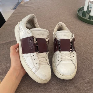 Valentino Garavani sneakers i vitt och vinrött - Snygga Valentino Garavani sneakers i vitt med vinröda detaljer. Skorna har en klassisk design med snörning och en bred vinröd rem över ovansidan. Perfekta för en stilren look.