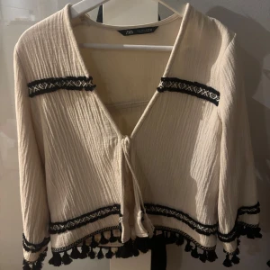 Beige cardigan jacka från Zara - Säljer en stilren beige cardigan jacka från Zara med svarta broderade detaljer och knyt framtill. Jackan har långa ärmar och dekorativa fransar längs nederkanten och ärmsluten. Perfekt för en bohemisk look!