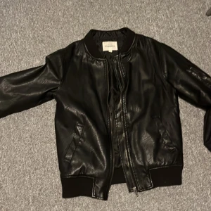 Svart skinnjacka från KappAhl 146 - Snygg svart skinnjacka från KappAhl med dragkedja framtill. Jackan har en klassisk bomberstil med ribbade muddar vid ärmslut och nederkant. Perfekt för en cool look! Super ifall du vill ge en bra present utan att förstöra hela plånboken. (Priset är inte spikat i sten)