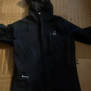 Svart Gore-Tex jacka från Haglöfs - Säljer en svart Gore-Tex jacka från Haglöfs. Jackan har en justerbar huva och dragkedjor framtill. Perfekt för utomhusaktiviteter med sitt vattentäta material och stilrena design. Passar bra för både regniga och blåsiga dagar.