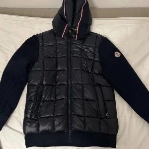 Svart quiltad jacka med huva - Snygg svart quiltad jacka med huva och dragkedja. Jackan har långa ärmar och en broderad logga på ärmen. Perfekt för kyligare dagar.