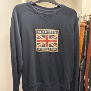 Säljer en stilren mörkblå tröja från Morris med ett stort tryck av Union Jack och texten 'Morris Since 1968' på framsidan. Tröjan har långa ärmar och en rund halsringning. Perfekt för en klassisk och avslappnad look.