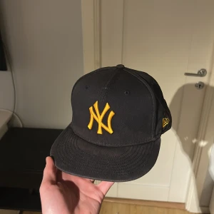 Mörkblå keps från New Era - Snygg mörkblå keps från New Era med gul broderad logga framtill. Lite missfärgning på böjen men annars super. Bra passform och är perfekt för dig som behöver en snygg keps.