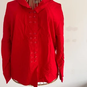 Röd blus från Lindex - Säljer en snygg röd blus från Lindex med dubbelknäppning framtill. Blusen har långa ärmar och en klassisk krage. Perfekt för att liva upp garderoben med en färgklick! 🔥Stl 38