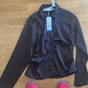 Brun blus från Samsøe Samsøe - Elegant brun blus från Samsøe Samsøe med långa ärmar och knappar framtill. Blusen har en stilren design med en lätt glansig yta och rynkdetaljer längs knappraden. Perfekt för en chic och sofistikerad look.