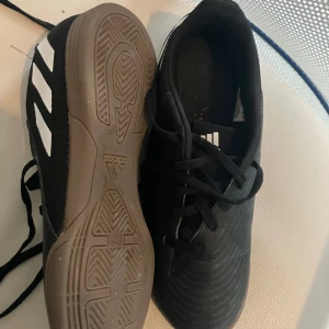 Svarta fotbollsskor från Adidas predator - Säljer ett par svarta fotbollsskor från Adidas med vita detaljer och snörning. Skorna har en klassisk design med tre vita ränder på sidan och en slitstark sula. Perfekta för inomhusfotboll eller träning.Använda ett fåtal gånger då jag köpte dem sent på futsalsäsongen.