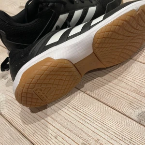 Svarta och vita sneakers från Adidas - Snygga svarta sneakers med vita ränder från Adidas. Skorna har en gummisula i ljusbrun färg och snörning för bra passform. Perfekta för sporter och träning.