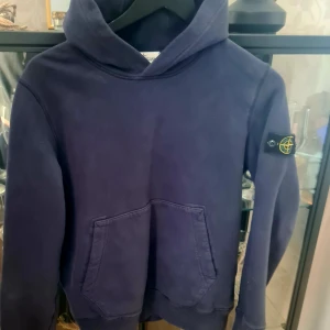 Mörkblå hoodie från Stone Island Junior - Säljer en snygg mörkblå hoodie från Stone Island Junior. Den har en klassisk känguruficka och det ikoniska märket på ärmen. Perfekt för en stilren och avslappnad look.,,junior i stl 146/152