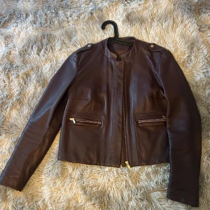 Brun/vinröd skinnjacka från Massimo Dutti - Snygg brun/vinröd skinnjacka från Massimo Dutti med dragkedja och stilrena detaljer. Jackan har långa ärmar och två framfickor med dragkedjor. Perfekt för en trendig look!