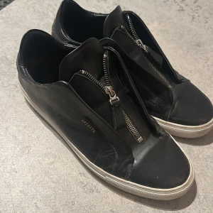 Svarta läder arigatos - Snygga svarta sneakers från Axel Arigato med dragkedja framtill. Bra skick men något år på ryggen och finns tyvärr ingen låda till. Jag säljer dessa skor för att det inte riktigt är min stil längre. Skriv privat för mer bilder och priset kan diskuteras vid snabbköp!
