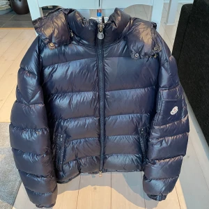 Moncler Maya - Säljer en snygg svart dunjacka från Moncler med dragkedja och huva. Jackan har en quiltad design och två praktiska fickor med dragkedja framtill. Perfekt för kalla dagar! Fint skick, ej använd ofta. Storlek 5, skulle säga att den passar som L ungefär. Enda slitningen är att spännena i luvan lossnat.