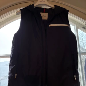 Svart dunväst från Moncler - Snygg svart dunväst från Moncler med dragkedja och huva. Västen har praktiska fickor med dragkedjor och en stilren design. Perfekt för kyligare dagar. Den har ett håll i högra fickan, den är storlek 14 år