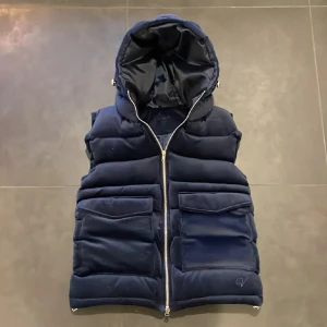 Vendiera väst - Snygg mörkblå dunväst med dragkedja och stor huva. Västen har två stora fickor framtill och är perfekt för kyligare dagar. Den quiltade designen ger en modern look.