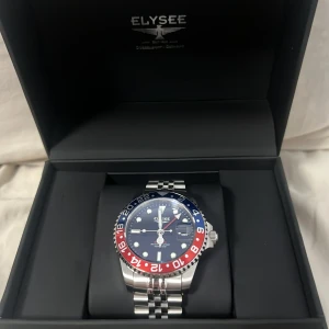 Elysee klocka med blå och röd bezel - Snygg Elysee klocka med en iögonfallande blå och röd bezel. Klockan har en svart urtavla med tydliga markeringar och ett datumfönster. Armbandet är i silverfärgat stål med en klassisk länkdesign. Perfekt för den som vill ha en stilren och sportig look.