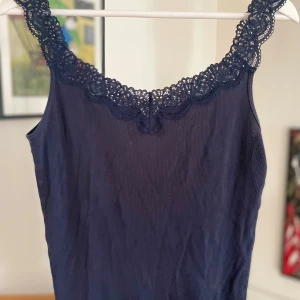 Mörkblå ribbad linnetopp med spetsdetaljer - Säljer en snygg mörkblå ribbad linnetopp med vackra spetsdetaljer runt halsringningen. Perfekt för sommardagar eller som ett lager under en kavaj. Toppen är ärmlös och har en tight passform som framhäver figuren. Ett måste i garderoben för en stilren look! Den sitter lite löst på mig som egentligen har strl xs
