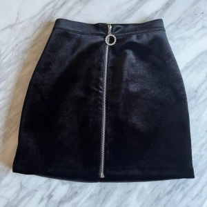 Svart sammetskjol med dragkedja - JätteSnygg svart sammetskjol med en dragkedja framtill. Kjolen har en kort längd och en elegant ringdetalj vid dragkedjan. Perfekt för en stilren look.