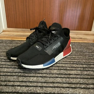 Adidas NMD - Snygga svarta Adidas NMD med färgglada detaljer i rött och blått. Skorna har en vit sula och klassisk snörning. Perfekta för en stilren och modern look. Använt de ett fåtal gånger