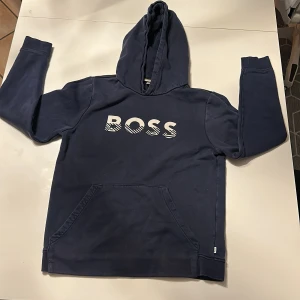 Mörkblå hoodie från BOSS - Säljer en snygg mörkblå hoodie från BOSS med stor logga på framsidan. Tröjan har en klassisk känguruficka och en bekväm huva. Perfekt för en avslappnad stil.