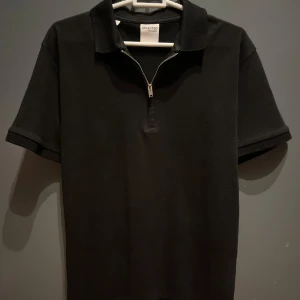 Svart pikétröja med half zip från Selected Homme - Snygg svart pikétröja från Selected Homme med dragkedja framtill. Perfekt för en stilren look. Använd 2 gånger säljer för den är för liten.