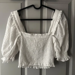 Vit blus med volanger - Säljer en söt vit blus med volanger och puffiga trekvartsärmar. Blusen har en smockad design som ger en fin passform och en romantisk känsla. 
