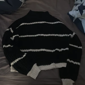 Svart och vit randig stickad tröja - Säljer en mysig svart stickad tröja med vita ränder. Tröjan har långa ärmar och en hög krage, perfekt för kyliga dagar. Den har en avslappnad passform och ribbade muddar vid ärmslut och nederkant. Den är använd ett få tal gånger. 