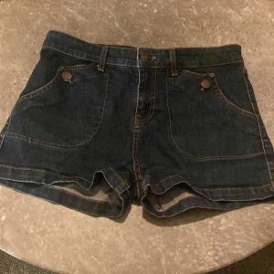 Mörkblå jeansshorts - Snygga mörkblå jeansshorts med knappdetaljer och framfickor. Shortsen har en klassisk design med två bakfickor och bälteshällor. Perfekta för en avslappnad stil. 
