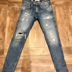 Blå jeans från replay - Säljer ett par blå jeans från replay med slim fit. De har en sliten look med flera revor och slitningar. Perfekta för en avslappnad stil. Midjan är normal och byxorna har klassiska fem fickor. Knappt använda nyskick
