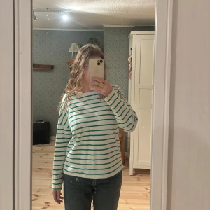 Randig långärmad tröja - Säljer en söt randig tröja med gröna och vita ränder. Tröjan har långa ärmar och en avslappnad passform, perfekt för en casual look. Den är också väldigt bekväm i materialet! Passar bra till jeans eller kjol. Köpt för 350kr