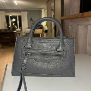 Grå väska  - Jättesöt grå väska som passar perfekt till att ha telefonen och läppglanset i på en utekväll. Den är rymlig, jag får plats med telefon, läppglans, plånbok, tuggummi och så vidare i den. Allt man behöver i en söt liten väska! Dragkedjan är sönder och den går inte att stänga. 