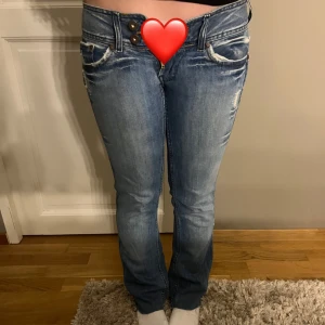 Pepe Jeans Victoria bootcut jeans - Snygga blå jeans från Pepe Jeans, modell Victoria. De har bootcut passform, låg midja och slitna detaljer framtill och bak. Jeansen har dubbla knappar i midjan och klassiska fickor. Perfekta för dig som gillar en avslappnad men trendig look.