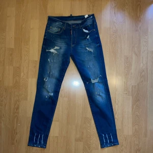 Dsquared2 blå skinny jeans herr - Snygga blå jeans från Dsquared2 i skinny fit med slitningar, färgstänk och distressed detaljer. Klassisk femficksmodell med coola slitna partier och målade färgprickar för en edgy vibe. Perfekta för dig som gillar streetstyle och vill sticka ut.