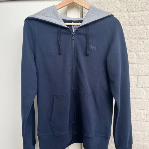 Mörkblå hoodie från Helly Hansen - Snygg mörkblå hoodie från Helly Hansen med hel dragkedja, grå insida på huvan och två stora fickor framtill. Klassisk logga på bröstet och justerbar snörning i huvan. Perfekt för chill dagar eller när du vill ha en enkel stil. Använt typ 4 gånger